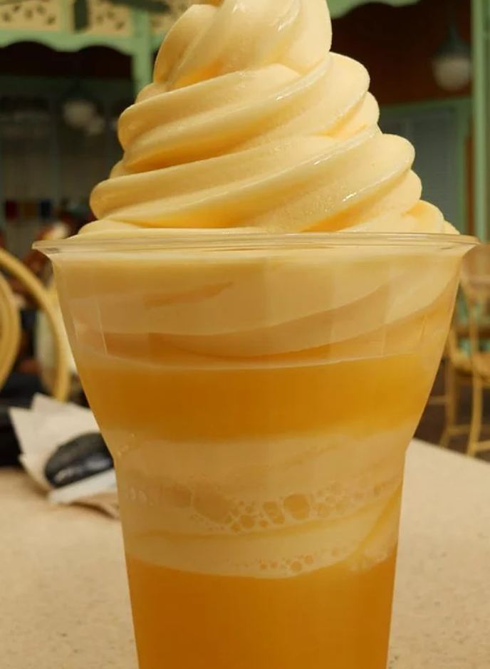 A close-up of a Dole Whip swirl in a waffle cone, topped with a maraschino cherry and a wedge of fresh pineapple.