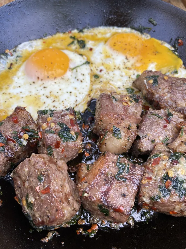 Ribeye steak bites covered in garlic-infused butter, served with sunny-side-up eggs on a plate.