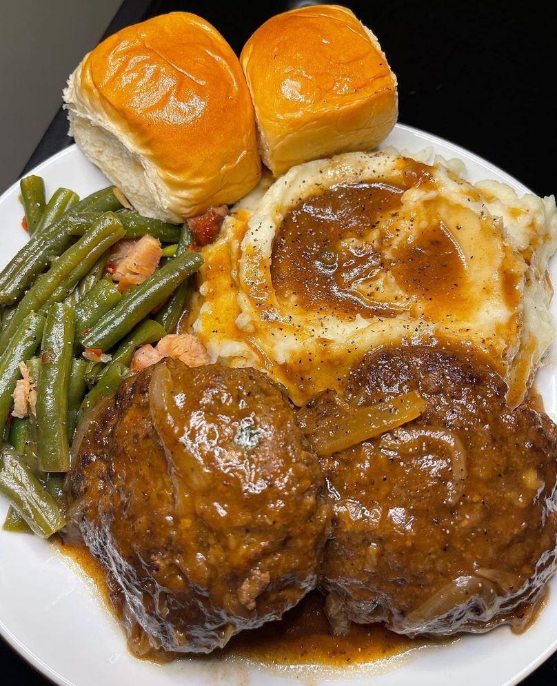 "A plate of Salisbury steak with onion gravy, mashed potatoes, string beans, and Hawaiian rolls"