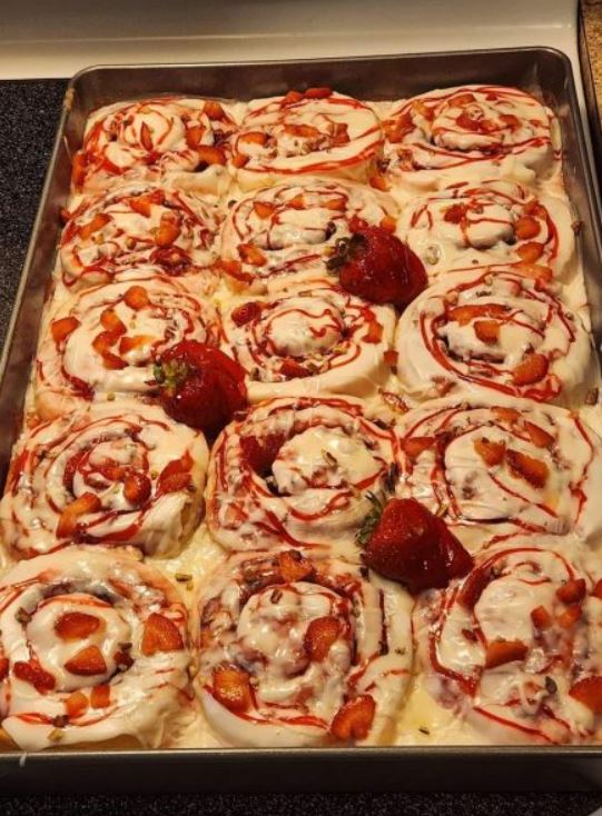 "A tray of freshly baked strawberry cheesecake cinnamon rolls with a sprinkle of pecans on top"