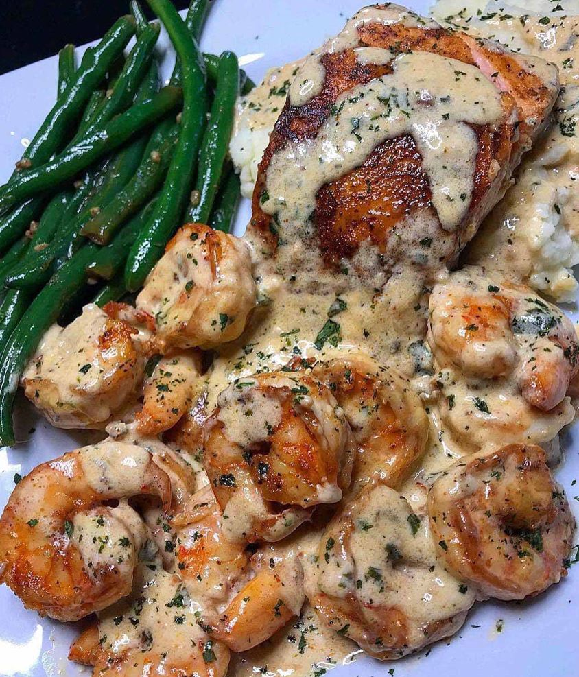 A plate of Cajun shrimp and salmon with garlic cream sauce, creamy mashed potatoes and garlic sautéed green beans. Perfect for a hearty and delicious meal.
