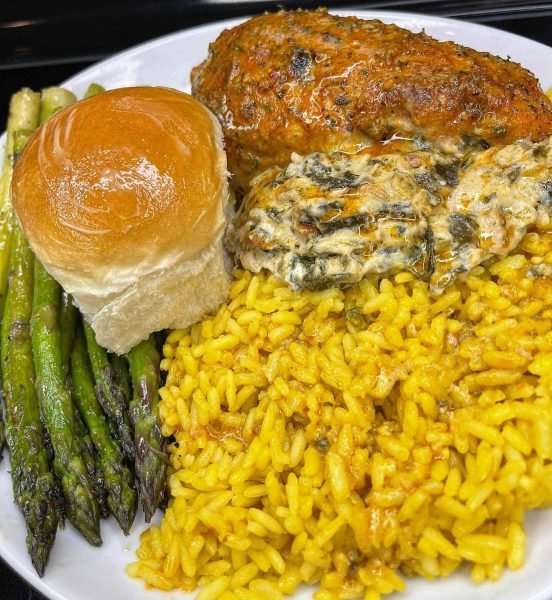 A plate of collard green dip stuffed chicken breast, yellow rice, asparagus, and a Hawaiian roll on a white plate.