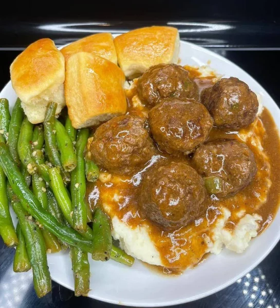 A close-up of a plate with meatballs and gravy over mashed potatoes, garnished with parsley and chives, with roasted green beans on the side.