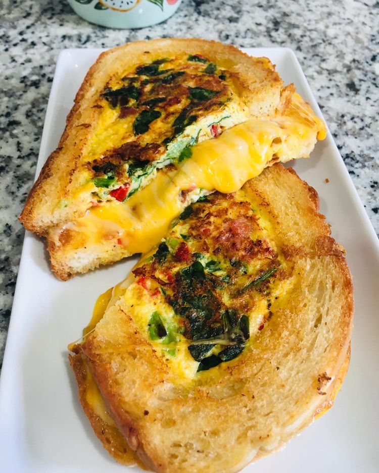 Close-up photo of a golden-brown grilled cheese sandwich cut in half, revealing a fluffy omelette filling with melted cheese and vegetables.