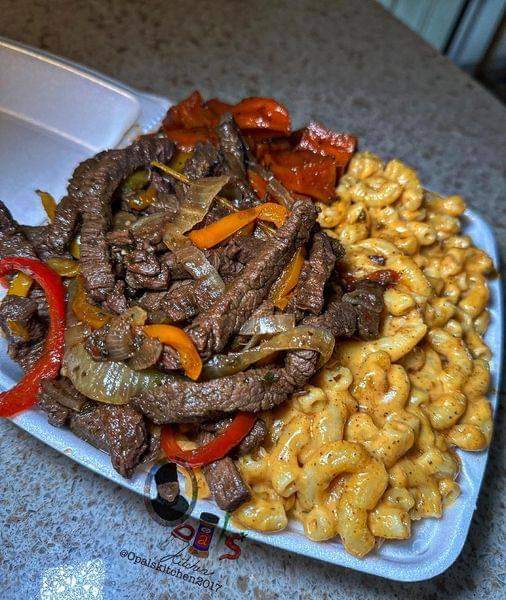 A photo of sizzling pepper steak pieces on a plate next to a bowl of creamy mac and cheese and roasted sweet potato wedges.