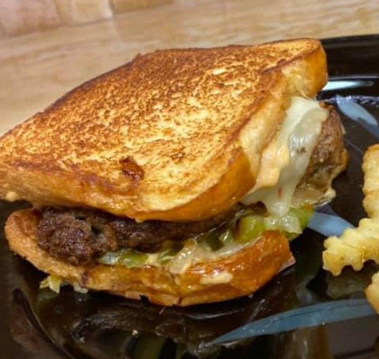 A close-up photo of a patty melt sandwich on a toasted bun. The sandwich is filled with a juicy hamburger patty, melted cheese, grilled onions, and a dollop of creamy secret sauce dripping down the side.