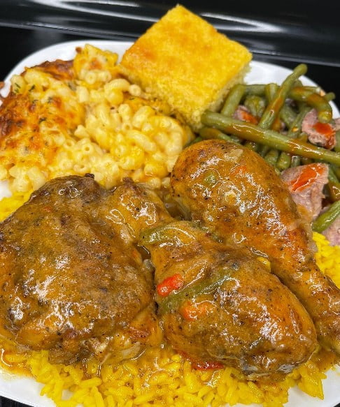 Smothered chicken pieces in a savory gravy on a bed of fluffy white rice, surrounded by baked mac and cheese, green beans with smoked turkey, and a slice of cornbread.