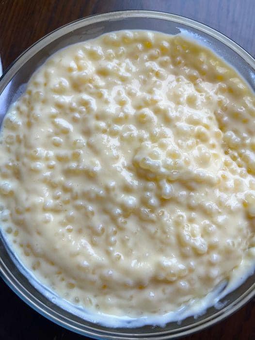 A close-up view of a glass bowl filled with homemade tapioca pudding. The pudding is topped with whipped cream and a maraschino cherry.